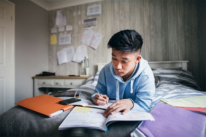Asian student on their bed doing homework in a notebook