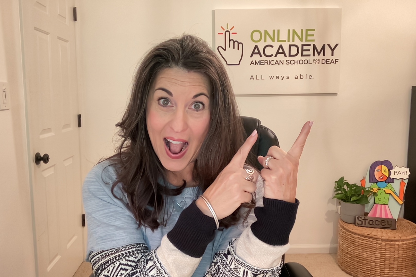 Enthusiastic white woman in a multi-patterned top and long dark hair, pointing toward a sign behind her saying "Online Academic American School for the Deaf ALL ways able." with ASD's hand logo on it