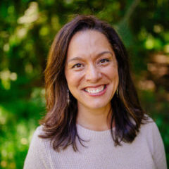 Smiling woman with olive skin wearing lipstick with dark shoulder-length hair. She is wearing a beige sweatshirt.