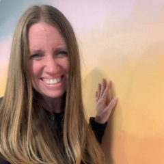 Smiling white woman with long brown hair, wearing a long-sleeved black top, standing by a pastel gradient wall with her hand on it