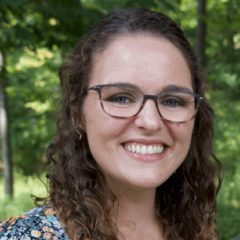 Smiling woman with glasses and curly shoulder-length hair in front of trees