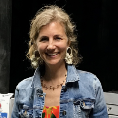 Smiling white woman in a denim jacket, colorful blouse and dangling earrings