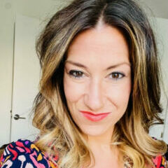 Smiling white woman with red lipstick and parted dark ombre shoulder-length hair. She is wearing a multi-patterned shirt.